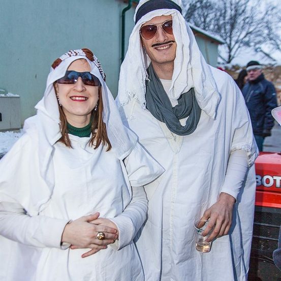 Ein Mann und eine Frau in weißen Roben und Sonnenbrillen stehen vor einem Gebäude. Der Mann hält ein Getränk in der Hand.