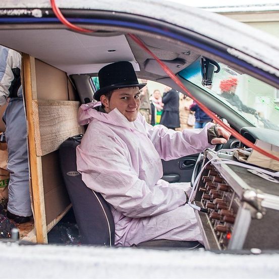Ein Mann in einem rosa Mantel und einem schwarzen Zylinder sitzt in einem Auto, wahrscheinlich für ein Fotoshooting. Er hält das Lenkrad mit der rechten Hand.