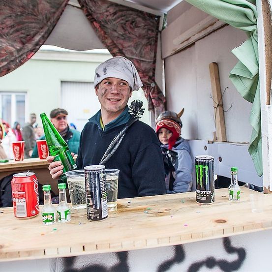 Ein Mann mit Bandana und Hörnerhelm steht hinter einem provisorischen Bartresen, hält eine grüne Flasche und ist von verschiedenen Getränken umgeben.