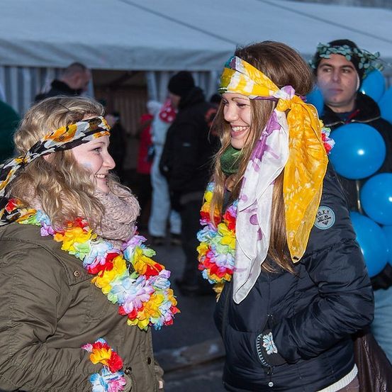 Zwei Frauen mit bunten Blumengirlanden und Kopftüchern lächeln sich bei einer Veranstaltung im Freien an. Dahinter hält ein Mann blaue Luftballons und eine Frau in einer grünen Jacke steht in der Nähe eines Zeltes.