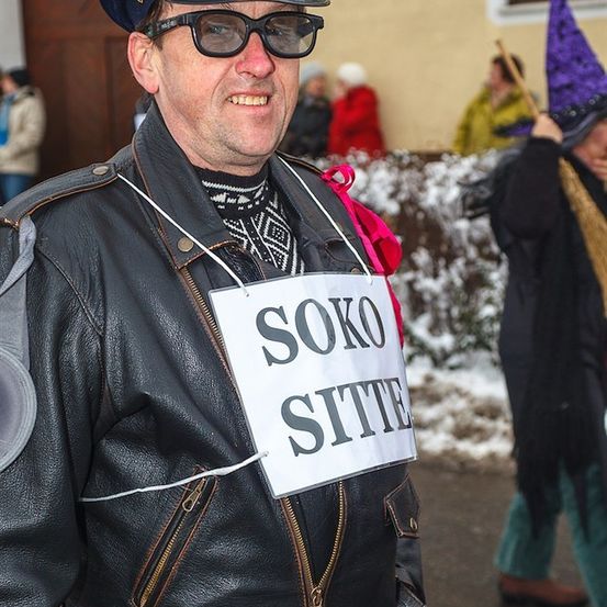 Ein Mann in einer Lederjacke mit einem Schild, das 'SOKO SITT' besagt, lächelt in einer verschneiten Umgebung mit Leuten in Kostümen hinter ihm.