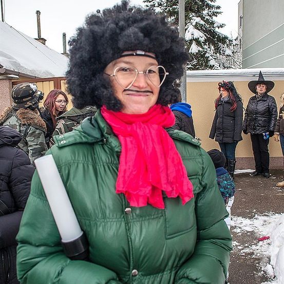 Eine Frau mit einer Afro-Frisur und einem roten Schal steht vor einem Gebäude und hält eine Taschenlampe. Hinter ihr sind Menschen in Winterkleidung, einer trägt einen Hexenhut.