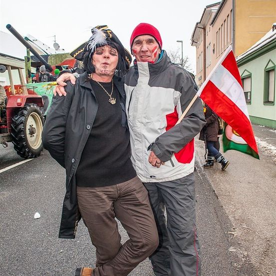 Zwei Männer in Kostümen, einer mit einem Piratenhut und der andere mit einer roten Mütze, stehen zusammen auf einer Straße. Der Mann im Piratenhut hält eine Flagge. Dahinter steht ein Traktor und ein Gebäude.
