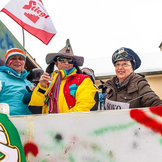 Drei Frauen in bunter Winterkleidung stehen hinter einer Absperrung, eine hält ein Glas, eine andere trägt einen Hut mit Brille, und eine Fahne mit dem Wort 'Stiegl' ist sichtbar.