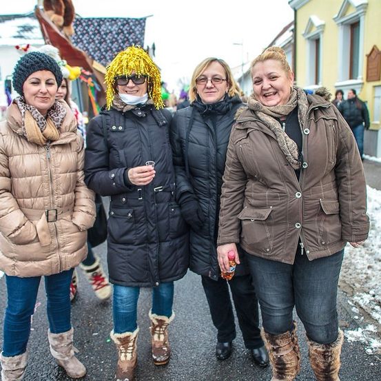 Vier Frauen in Wintermänteln und Stiefeln stehen auf einer verschneiten Straße und lächeln für die Kamera. Eine hält ein Getränk und sie sind von festlichen Dekorationen und Gebäuden umgeben.