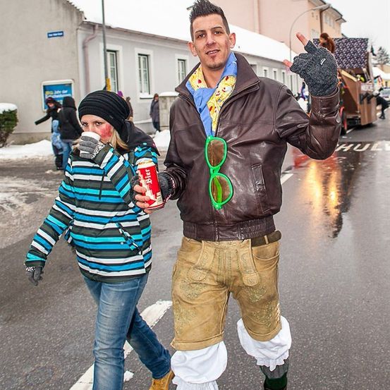 Ein Mann in einer braunen Lederjacke und grünen Fliegerbrille posiert für ein Foto mit einer Frau in einer gestreiften Jacke, beide gehen auf einer verschneiten Straße.