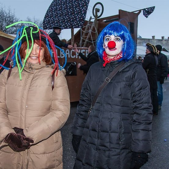 Zwei Frauen in Winterkleidung, eine mit blauen Haaren und einer roten Clownsnase, die andere mit bunten Haarverlängerungen, stehen bei einer Outdoor-Veranstaltung.