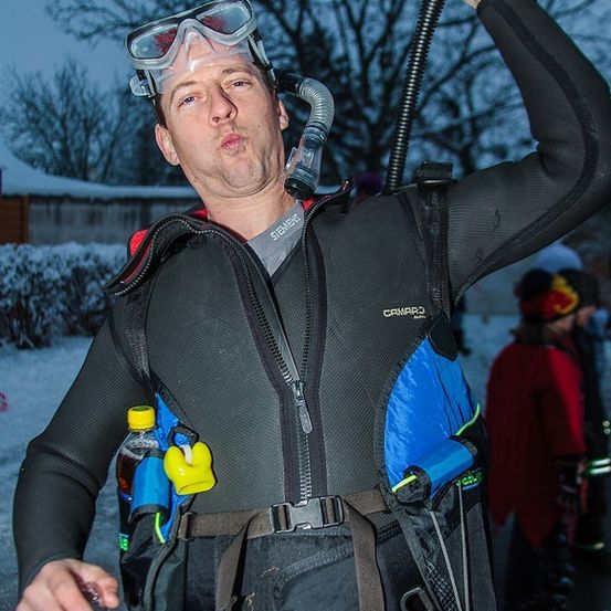 Ein Mann in einem schwarzen und blauen Neoprenanzug mit Schnorchel und einer gelben Flasche am Brustgurt steht in einem verschneiten Bereich.