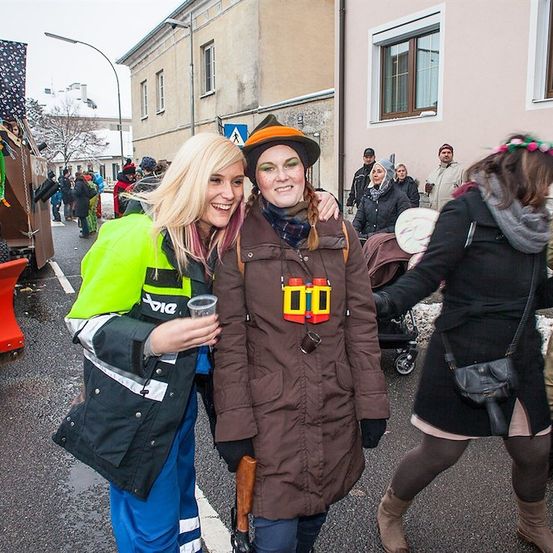 Zwei Frauen in festlicher Kleidung, eine in einer reflektierenden Sicherheitsjacke und die andere in einem braunen Mantel, posieren für ein Foto auf einer verschneiten Straße.
