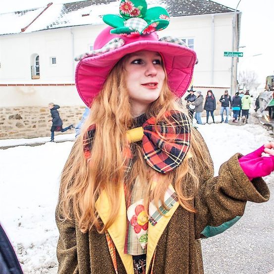 Eine Frau in einem skurrilen Kostüm steht im Schnee, trägt einen rosa Hut mit Blumen und eine karierte Schleife. Hinter ihr gehen Menschen auf einer verschneiten Straße.