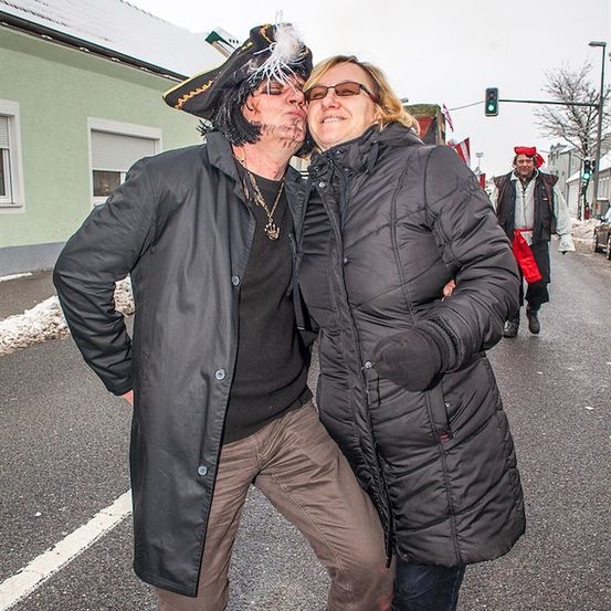 Ein Mann und eine Frau in Winterkleidung und Piratenkostümen stehen in der Mitte der Straße, lächeln und posieren für ein Foto. Hinter ihnen geht ein Mann in Piratenkostüm die Straße entlang.
