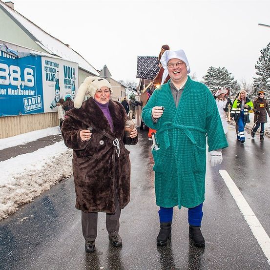 Zwei Menschen in Winterkleidung und Bademänteln gehen lächelnd und mit Getränken in der Hand eine verschneite Straße entlang. Dahinter eine Parade mit Menschen in Kostümen und einem Banner mit einem Radiosenderlogo.
