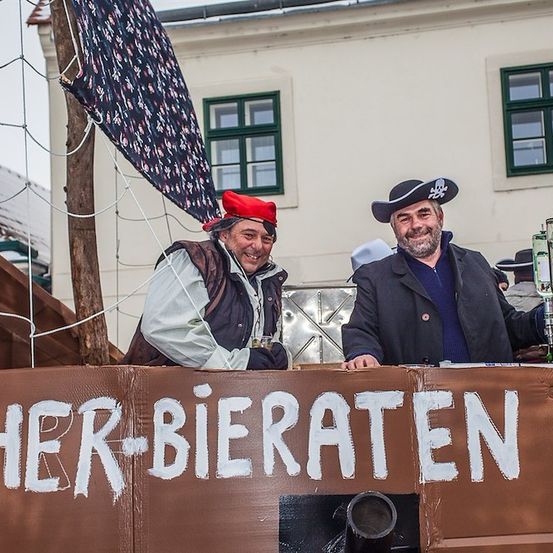 Zwei Männer in Piratenkostümen stehen auf einem Festwagen mit einem Schild, das 'HER-BIERRATEN' besagt. Der Mann auf der linken Seite trägt einen roten Hut und ein Augenklappe, während der Mann auf der rechten Seite einen schwarzen Hut trägt. Dahinter steht ein Gebäude mit grünen Fenstern.