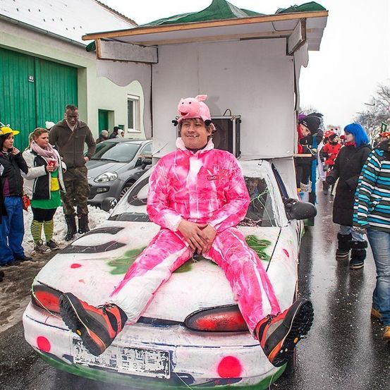 Eine Person, die als Schwein verkleidet ist, sitzt auf einem dekorierten Auto mit einer Schweinemaske, umgeben von einer Menge in einer festlichen Atmosphäre.