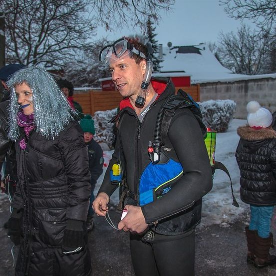 Ein Mann in einem Taucheranzug steht im Schnee mit einer Frau in einer silbernen Perücke. Sie sind von anderen Menschen umgeben, von denen einige Mützen und Stiefel tragen.