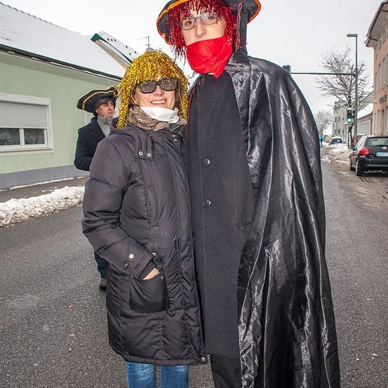 Eine Frau mit gelber Perücke und ein Mann mit rotem Maske und Umhang stehen auf einer verschneiten Straße. Dahinter geht ein Mann mit Piratenhut.