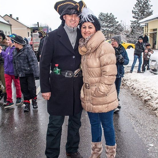 Ein Mann und eine Frau in Winterkleidung und Kostümen stehen im Schnee mit einer Gruppe von Menschen im Hintergrund.