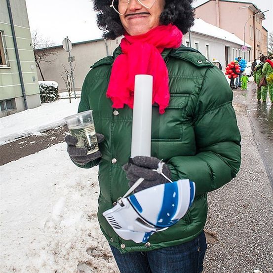 Eine Frau mit lockigem Haar, die eine grüne Jacke, einen roten Schal trägt und einen Plastikbecher und eine weiße Röhre auf einer verschneiten Straße hält.