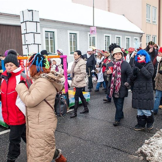 Eine Gruppe von Menschen in Winterkleidung und einigen in Kostümen geht bei einem Umzug auf einer verschneiten Straße. Einige halten Tassen und andere tragen Hüte und Gesichtsbemalung.