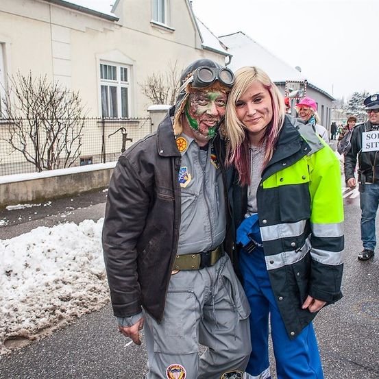 Ein Mann in einem Zombie-Kostüm und eine Frau in einer neonfarbenen Jacke stehen auf einer verschneiten Straße, mit einem Polizisten, der ein Schild in der Hand hält.