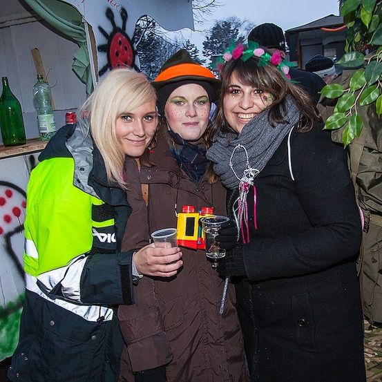 Drei Frauen in Winterkleidung, zwei mit Getränken, stehen vor einer mit Graffiti bemalten Wand. Die Frau links trägt eine neongrüne und schwarze Jacke.