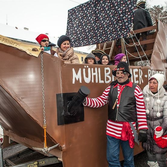 Eine Gruppe von Menschen in Kostümen, darunter ein Pirat, steht auf einem großen Pappboot während einer Parade. Das Boot hat ein Schild mit der Aufschrift 'Mühlbauer'.