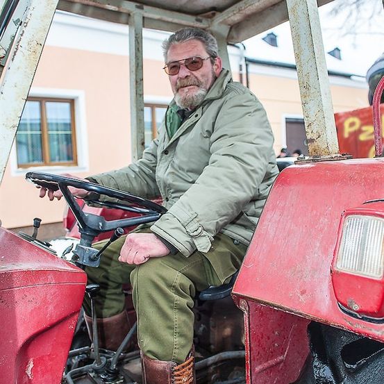 Ein älterer Mann mit Bart, der eine Brille und eine grüne Jacke trägt, sitzt auf einem roten Traktor.