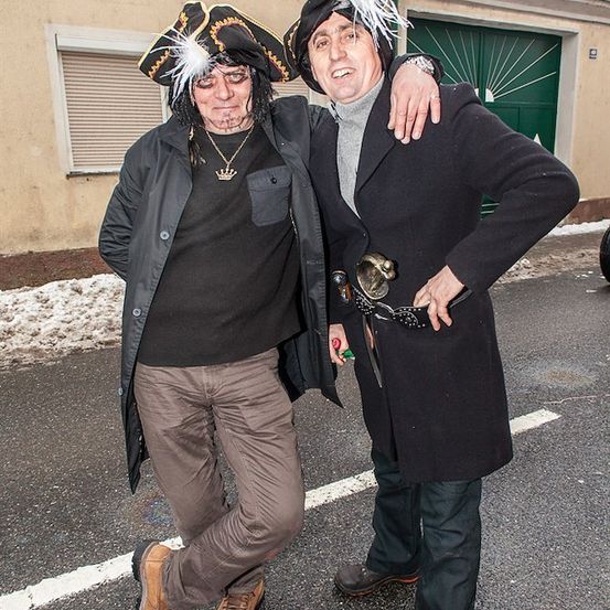 Zwei Männer in Piratenkostümen posieren für ein Foto auf einer verschneiten Straße. Einer trägt einen Piratenhut und eine Halskette, der andere hat einen Gürtel mit Schnalle.
