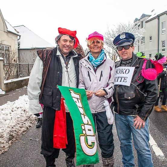 Drei Menschen in Kostümen stehen lächelnd für die Kamera auf einer verschneiten Straße. Der Mann links trägt eine Piratenhaube und ein rotes Bandana, die Frau in der Mitte trägt eine rosa Haube und einen Schal und der Mann rechts trägt eine Lederjacke und hält ein Schild.