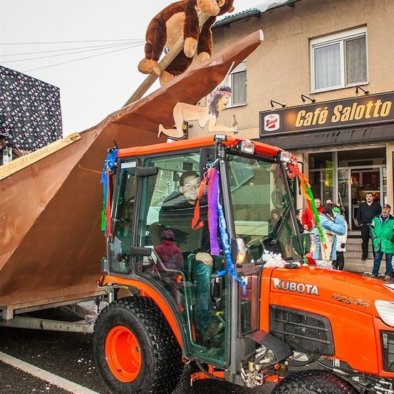 Ein dekorierter orangefarbener Kubota-Traktor mit einer großen braunen Plane und einer Affen- und Meerjungfrauenfigur darauf steht vor einem Gebäude mit einem Schild, das Cafe Salotto liest.