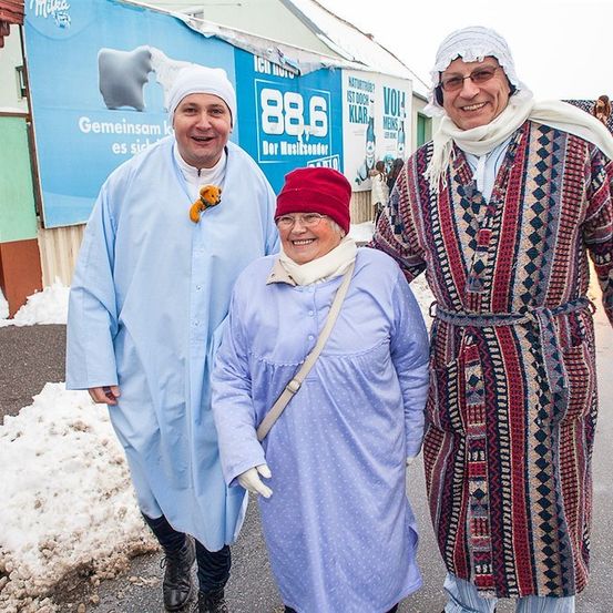 Drei Menschen in Winterkleidung stehen zusammen in einem verschneiten Bereich. Der Mann links trägt einen blauen Umhang, die Frau in der Mitte trägt ein blaues Kleid und einen roten Hut, und der Mann rechts trägt einen gemusterten Umhang. Hinter ihnen befindet sich ein blaues Banner mit Text und einem Bild eines Eisbären.
