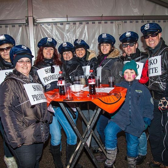 Eine Gruppe von Menschen in Polizeiuniformen, einige halten Schilder, und ein Kind vorne in einem grünen Hut und blauem Mantel. Sie stehen um einen Tisch mit Getränken und einem Banner.