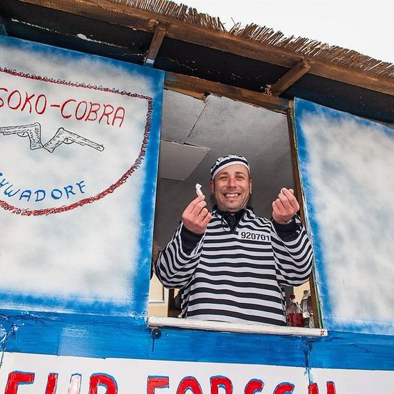 Ein Mann in einem gestreiften Shirt steht in einer Kabine mit einem Schild, das 'SOKO-COBRA SCHWADORF' besagt, und hält ein kleines Objekt in der Hand.