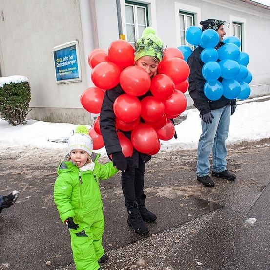 Eine Frau mit einem großen Strauß roter Luftballons geht mit einem Kind in einem grünen Schneeanzug auf einer verschneiten Straße.