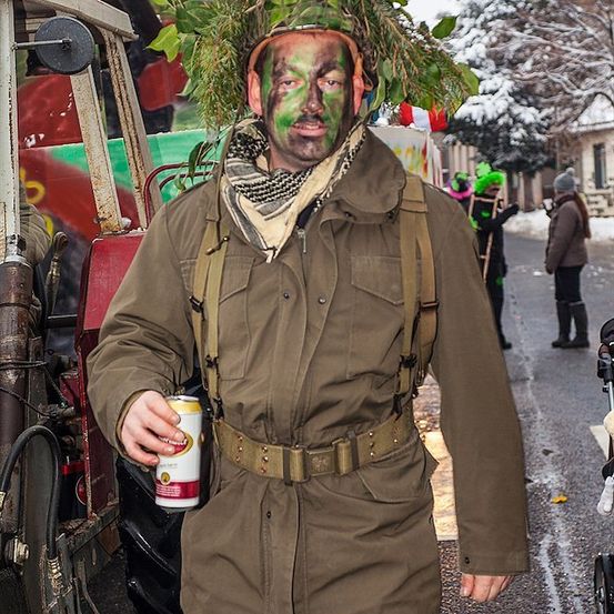 Ein Mann in einer Militäruniform mit grüner Gesichtsmaske und Blättern auf dem Kopf, der eine Bierdose hält.