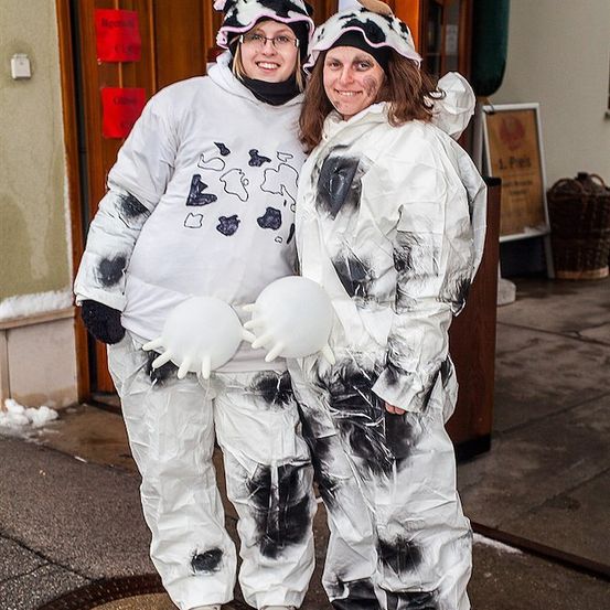 Zwei Frauen in Kuhkostümen, eine mit Hut und die andere mit Hörnern, stehen vor einem Gebäude mit schneebedecktem Boden.