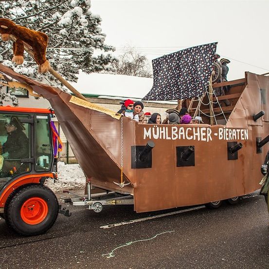 Ein Paradewagen in Form eines Piratenschiffs wird von einem Traktor auf der Straße gezogen. Es hat eine Besatzung von Menschen in Piratenkostümen und ein großes Affenspielzeug hängt vorne. Der Wagen ist mit einem schwarz-weiß gemusterten Segel dekoriert.