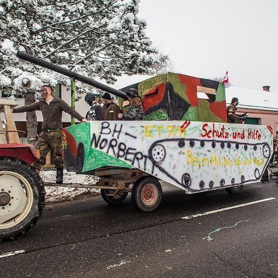 Eine Gruppe von Menschen in Militäruniformen fährt auf einem Traktor, der einen dekorierten Panzeranhänger mit Graffiti zieht. Der Panzer hat ein Tarnmuster und eine Flagge oben. Die Leute halten sich an den Seiten des Panzers fest. Im Hintergrund befinden sich Bäume und Gebäude.