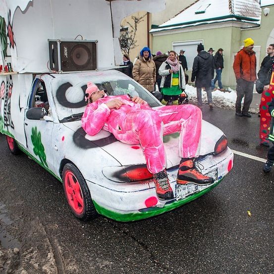 Ein Mann in rosa Overalls liegt auf einem dekorierten Auto, möglicherweise bei einem Winterfest, mit Leuten in farbenfrohen Kostümen um ihn herum.