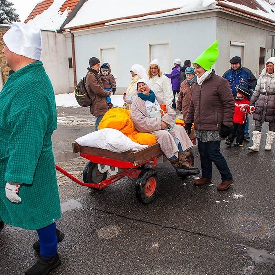 Eine Gruppe von Menschen in Winterkleidung und Mützen geht auf einer verschneiten Straße, wobei eine Person auf einem roten Wagen mit Kissen sitzt.