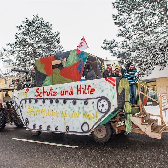 Ein dekorierter Traktor mit Leuten darauf, der auf einer verschneiten Straße fährt. Der Traktor ist bemalt, um wie ein Panzer mit Graffiti und einer roten Flagge auszusehen.