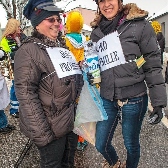 Zwei Frauen in Winterkleidung stehen auf einer verschneiten Straße, eine hält eine Milchflasche und ein Schild mit der Aufschrift 'SOKO PROUD', die andere lächelt und hält eine Plastiktüte. Hinter ihnen sind eine Person in einem gelben Kostüm und ein Trompeter zu sehen.