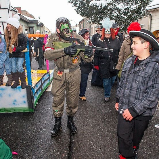 Eine Gruppe von Menschen in verschiedenen Kostümen, darunter ein Soldat, geht auf einer verschneiten Straße. Ein Kind in einem Eselskostüm ist auf einem Wagen, und ein Junge in einem Cowboyhut spricht mit einem Soldaten.