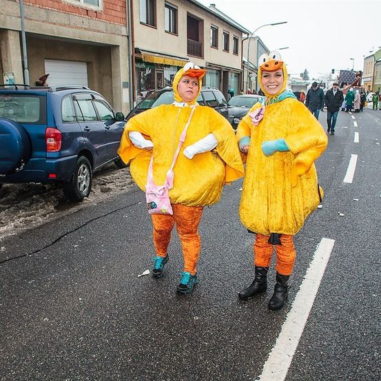 Zwei Frauen in gelben Entenkostümen stehen auf einer verschneiten Straße und lächeln für die Kamera. Dahinter ist ein blauer SUV geparkt, und Menschen gehen in der Ferne.