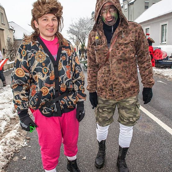 Zwei Personen in bunten und Tarnoutfits stehen auf einer verschneiten Straße. Die Frau hat ein pinkes Oberteil und pinke Hosen, während der Mann eine Tarnjacke und Tarnhosen trägt.