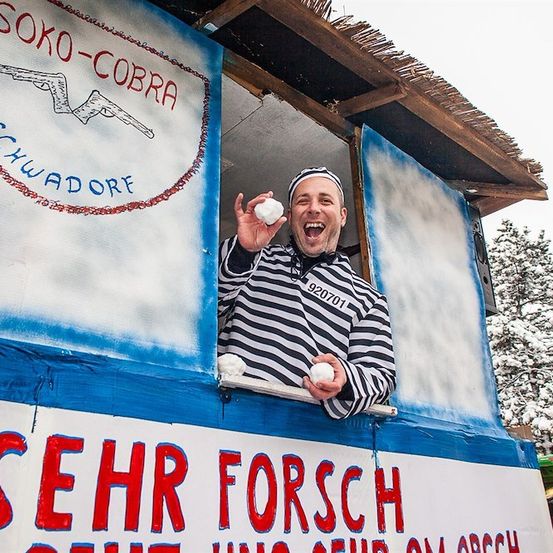 Ein Mann in einem gestreiften Shirt und einer Mütze wirft Schneebälle aus einem Stand mit dem Schild 'Sehr Forsch'. Der Stand hat ein Holzdach und einen blau-weißen Hintergrund. Der Mann lächelt und hält einen Schneeball in der Hand.