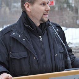 Ein Mann mit kurzem Haar und Bart trägt eine schwarze Jacke und steht an einem Podium mit einem Mikrofon vor ihm.