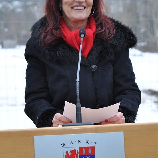 Eine Frau mit roten Haaren steht an einem Rednerpult, lächelt und hält ein Papier in der Hand, mit einem Mikrofon vor ihr. Sie trägt einen roten Schal und einen schwarzen Mantel mit Pelzbesatz. Hinter ihr ist ein verschneiter Hintergrund.