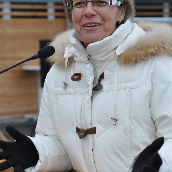 Eine Frau in einer weißen Jacke mit Pelzkragen und schwarzen Handschuhen lächelt und spricht in ein Mikrofon.