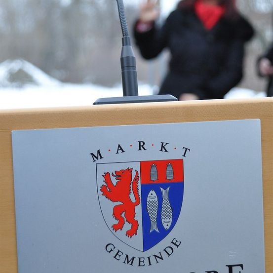Ein Podiumsschild für die Gemeinde Schwadorf mit einem Wappenschild, der einen roten Löwen und zwei Fische auf blauem Hintergrund zeigt. Eine Frau in einem schwarzen Mantel steht im Hintergrund.